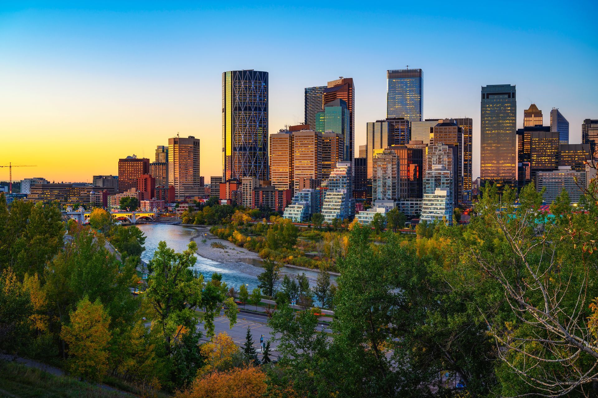 A city skyline with a river in the foreground and trees in the background.