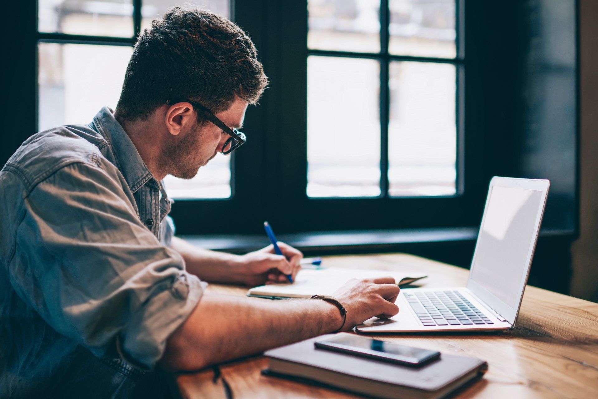A man is sitting at a table with a laptop and writing on a piece of paper.