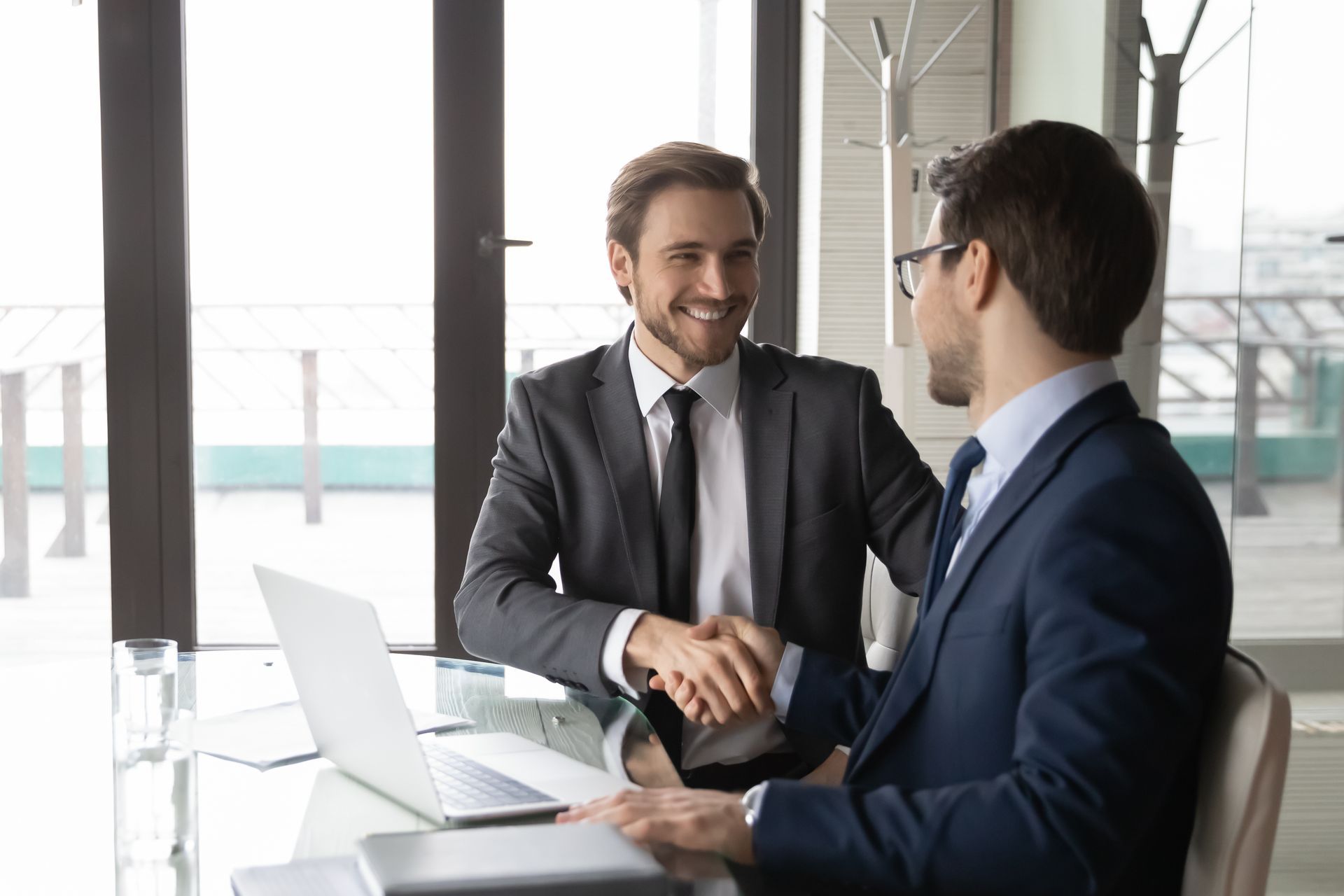 Two businessmen are shaking hands while sitting at a table with a laptop.