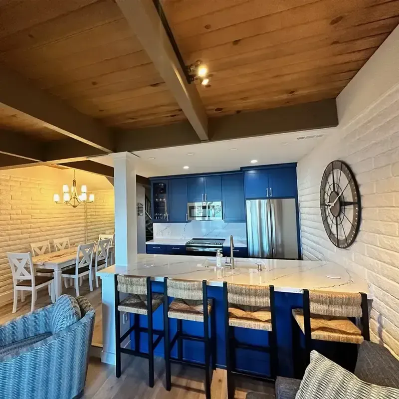 Open-concept kitchen with blue cabinets, a quartz island with four stools, and a dining area against a white brick wall.