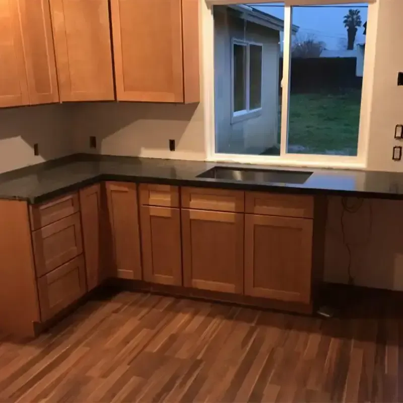 A kitchen interior with wooden cabinets, dark countertops, a sink under a window, and wood-patterned flooring.