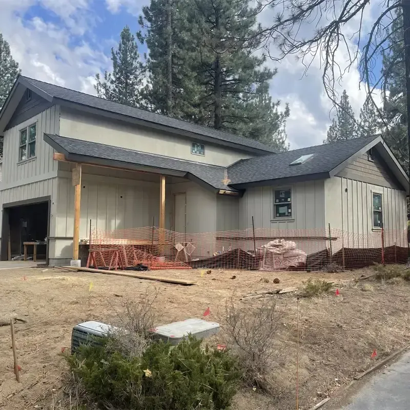A two-story house under construction with light-colored vertical siding, a dark roof, and an open front porch area.