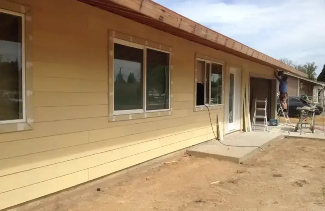 A person stands on a ladder working on the exterior siding of a light yellow house under a blue, cloudy sky.