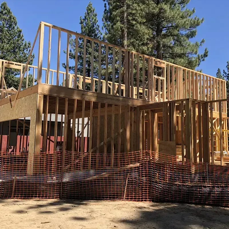 The wooden framing of a two-story residential house under construction, set against a backdrop of pine trees.