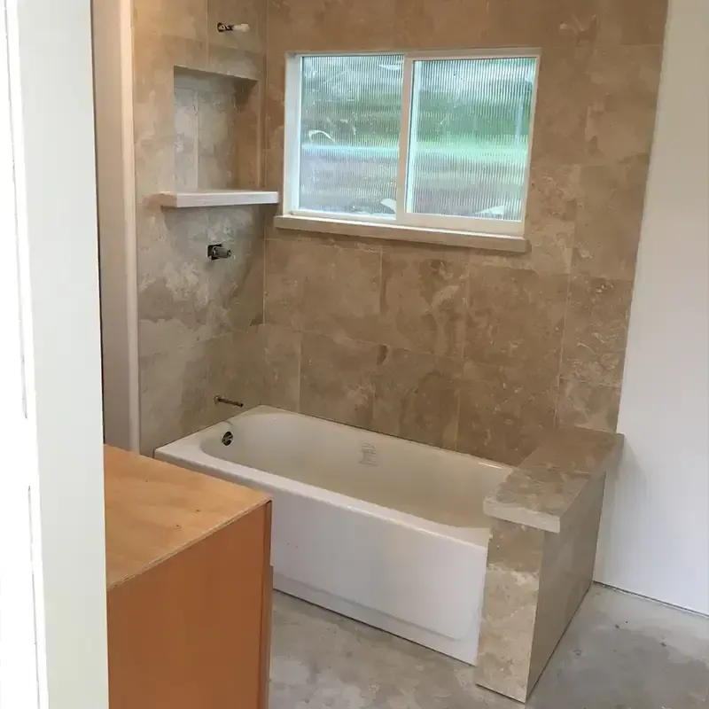 A bathroom under renovation featuring a white bathtub and beige tiled walls with a built-in recessed shelf and window.