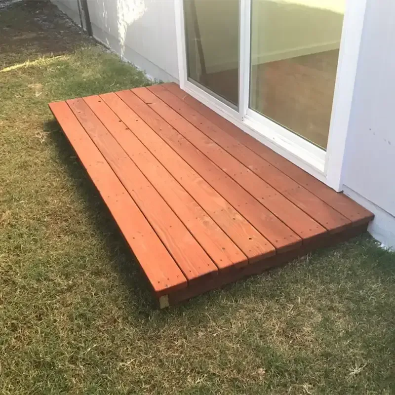 A small, rectangular deck made of stained reddish-brown wooden planks sits on a patch of grass outside a sliding glass door.