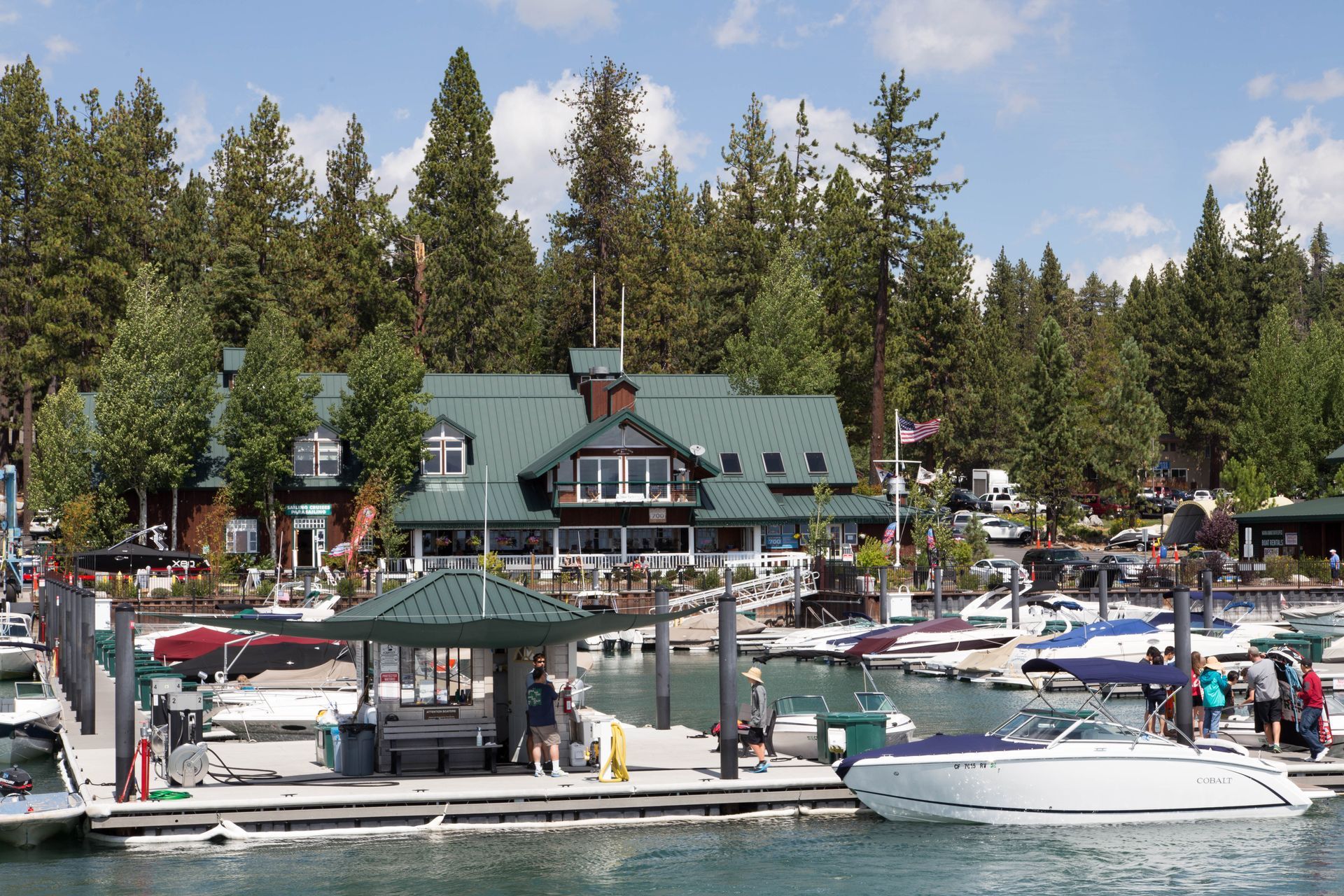 A green-roofed lodge sits on a tree-lined lakeshore behind a busy marina filled with docked motorboats.