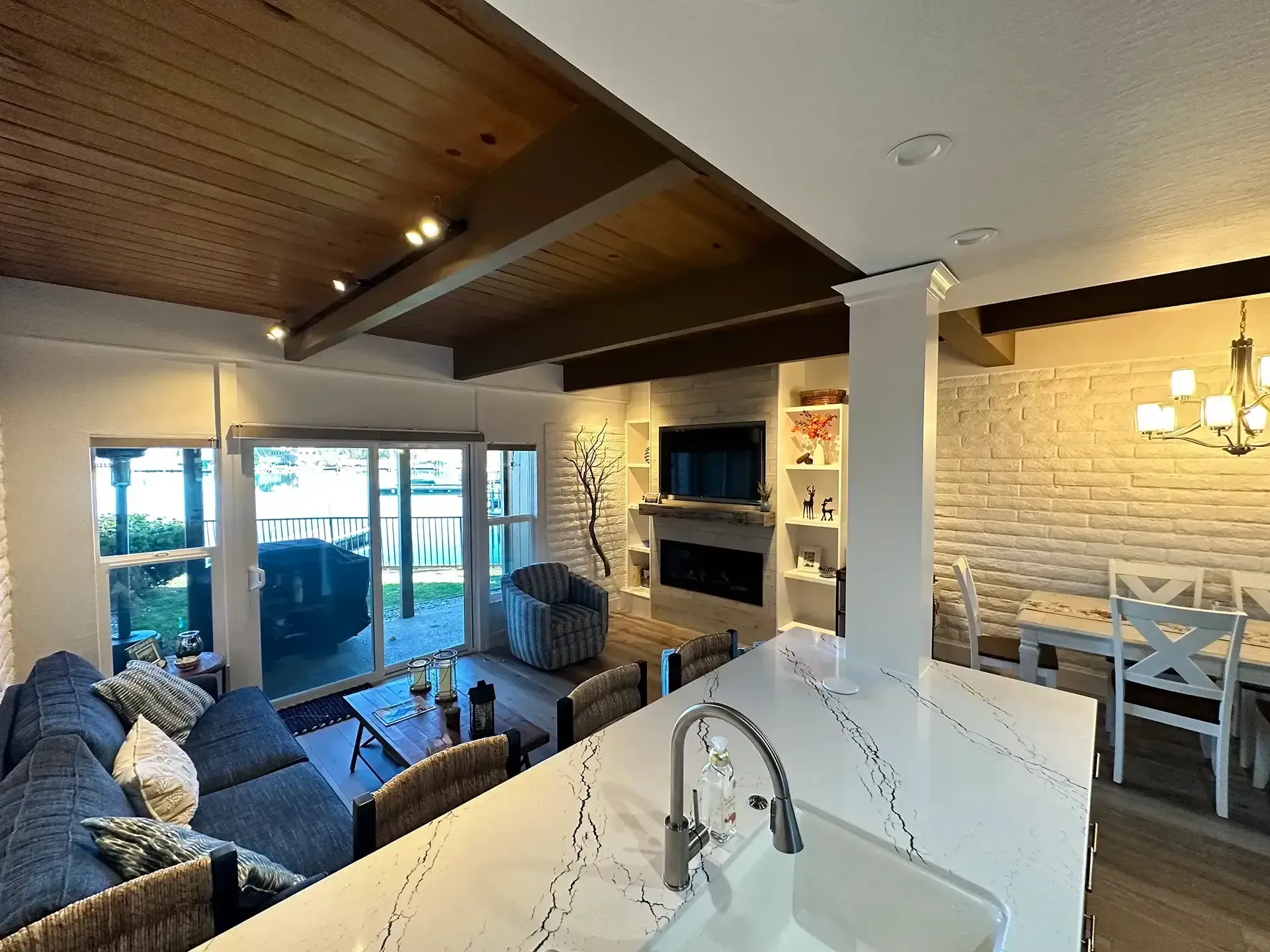 An open-concept living area with a blue sofa, modern kitchen island, fireplace, and dining table, featuring wood ceilings.