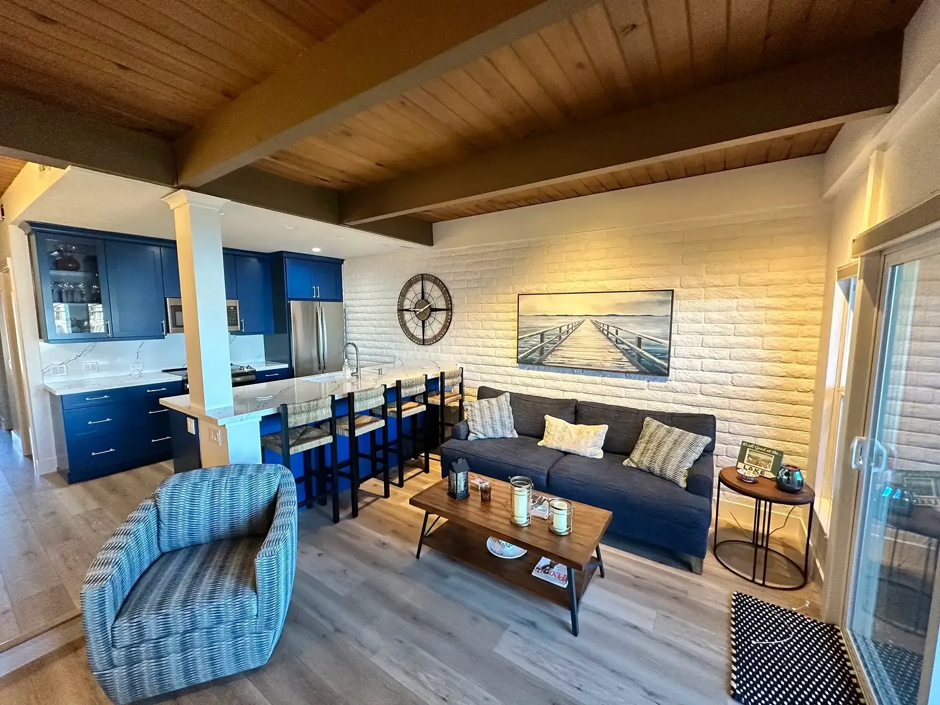 Open-plan living room with wood ceiling, white brick wall, blue sofa, patterned armchair, and a modern blue kitchen.