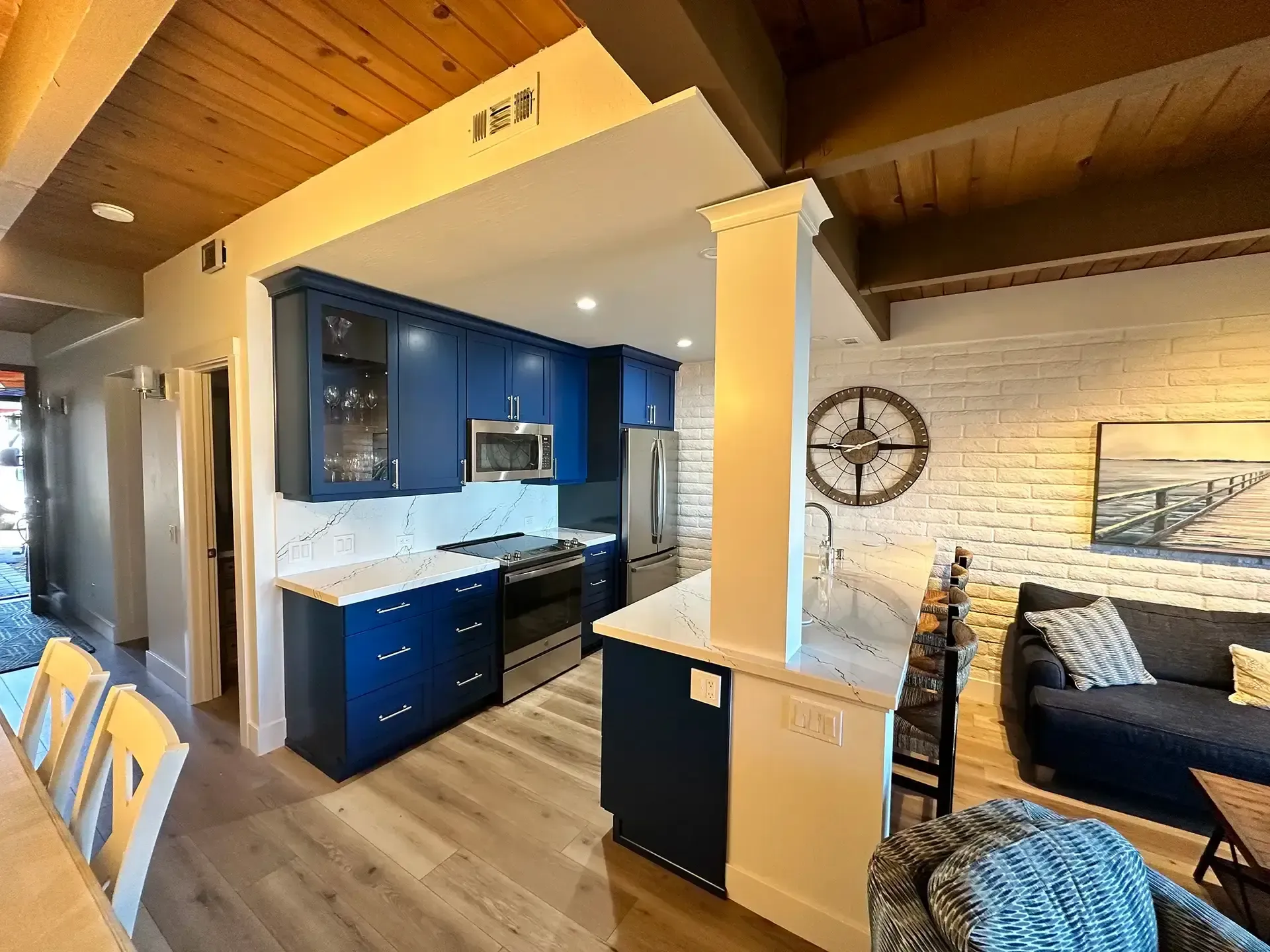 A modern kitchen with navy blue cabinets and white countertops, adjacent to a living area with a white brick wall.