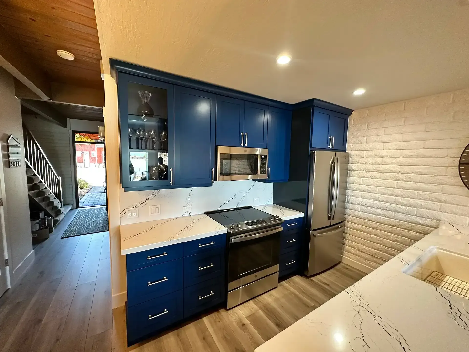 A kitchen featuring dark blue cabinets, white marble-patterned countertops, stainless steel appliances, and a brick wall.
