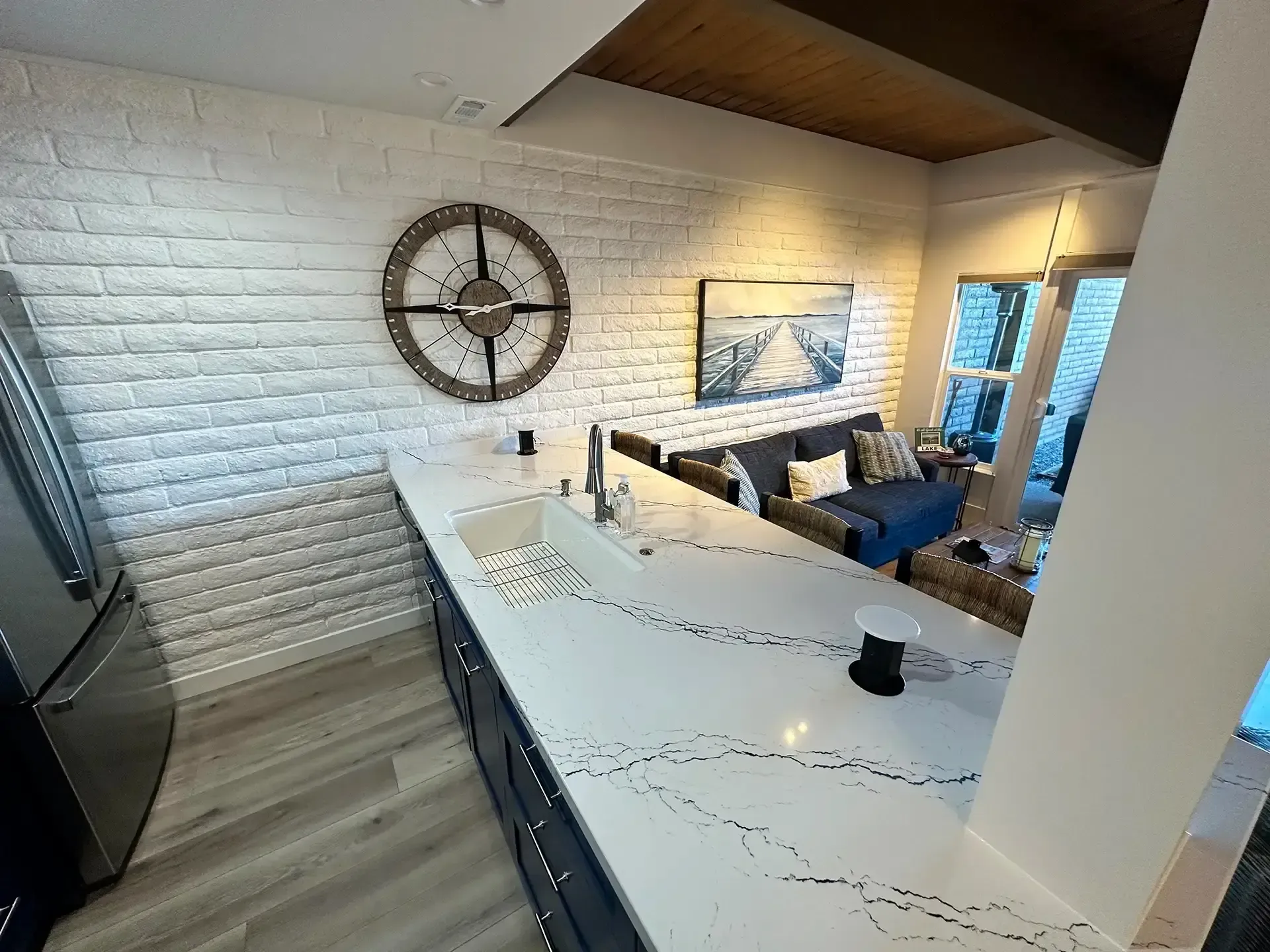 A modern kitchen with white marble countertops and dark cabinets, featuring a white brick wall with a large round clock.