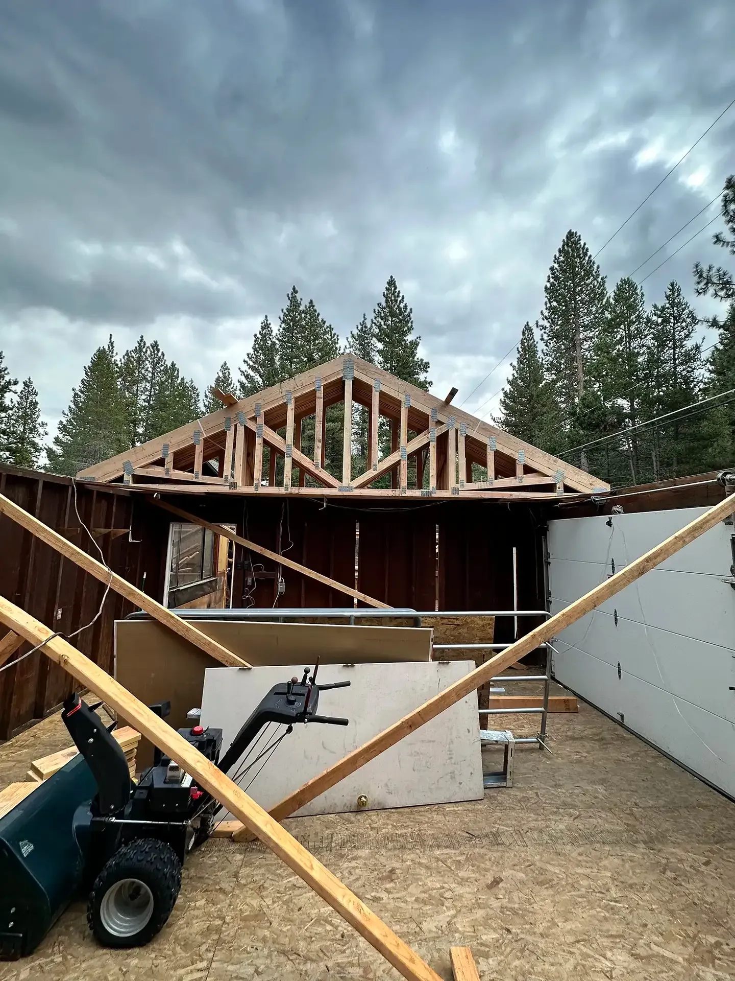 An unfinished wooden roof frame sits atop walls in an outdoor construction site with pine trees and a cloudy sky above.