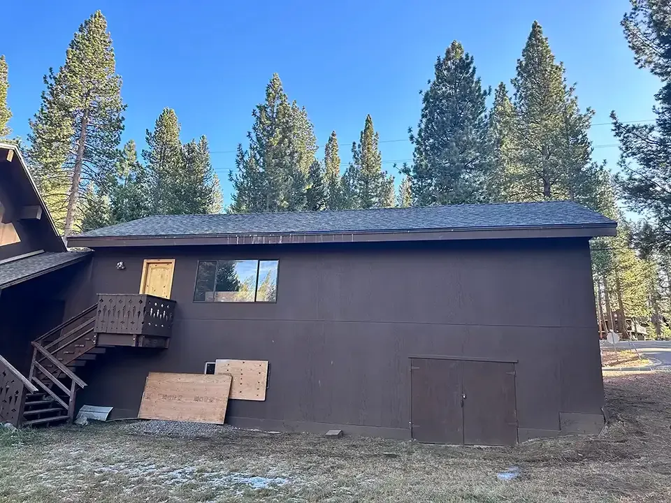 A dark brown two-story house with a wooden deck, a single window, and boarded-up areas, set against a forest background.
