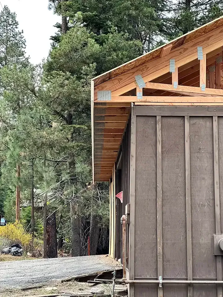 The side view of a building under construction, featuring exposed wooden roof trusses and brown siding in a forested area.