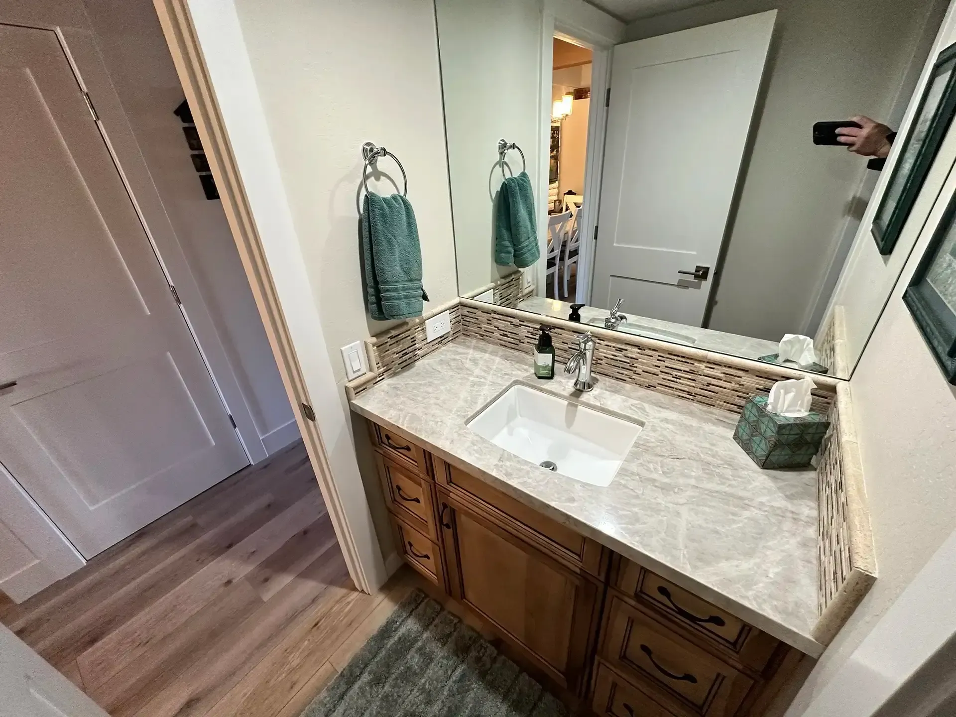 A bathroom vanity with a marble countertop, white sink, wooden cabinets, and a mirror reflecting a doorway.