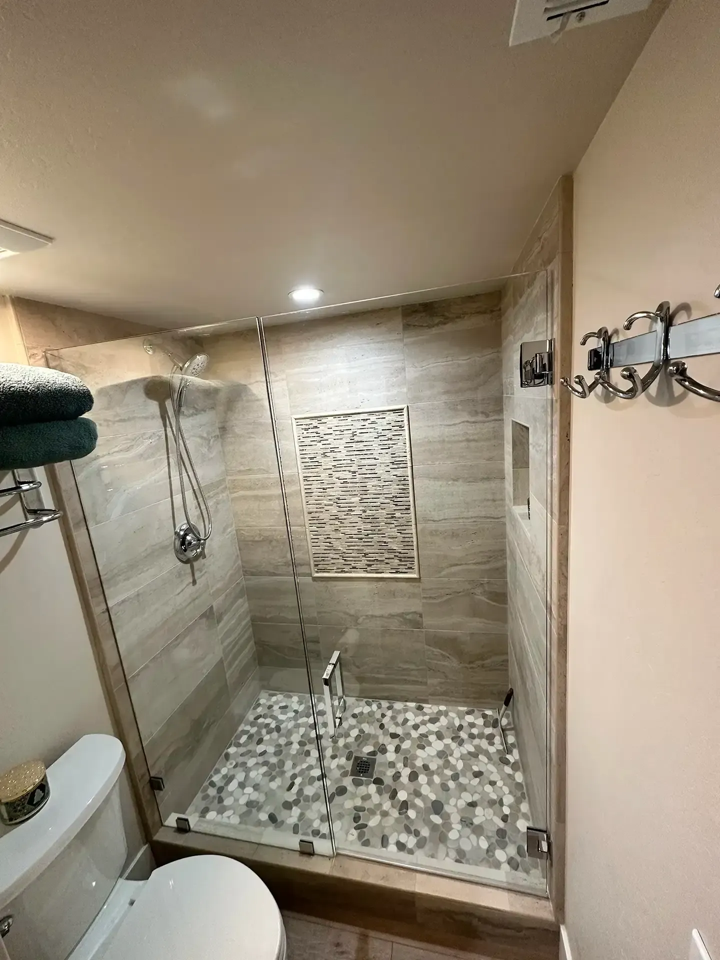 A glass-enclosed shower with beige tile walls, a pebble-stone floor, and an inset decorative tile panel.