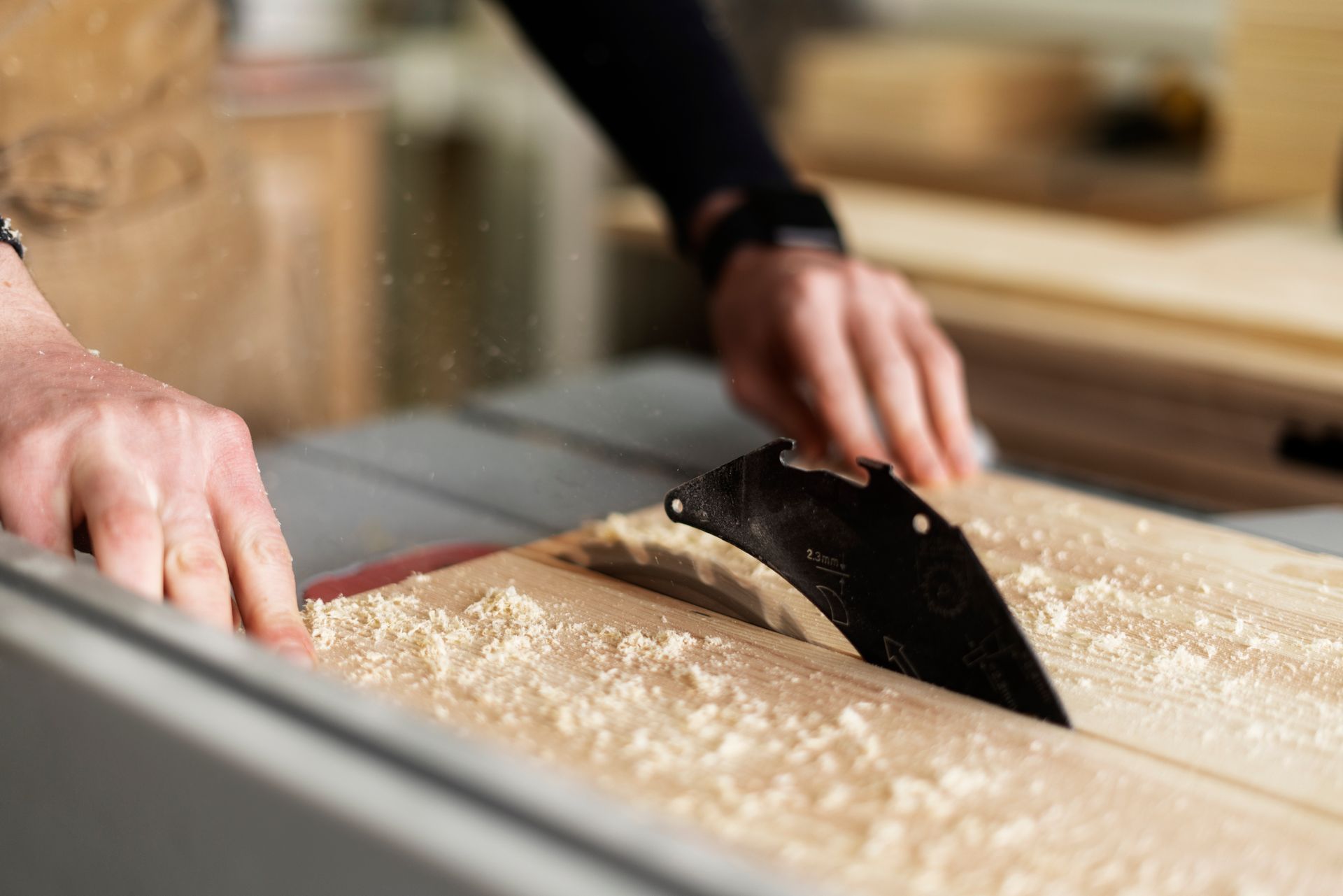 Hands pushing a wooden board through a table saw in a woodworking shop, creating sawdust.