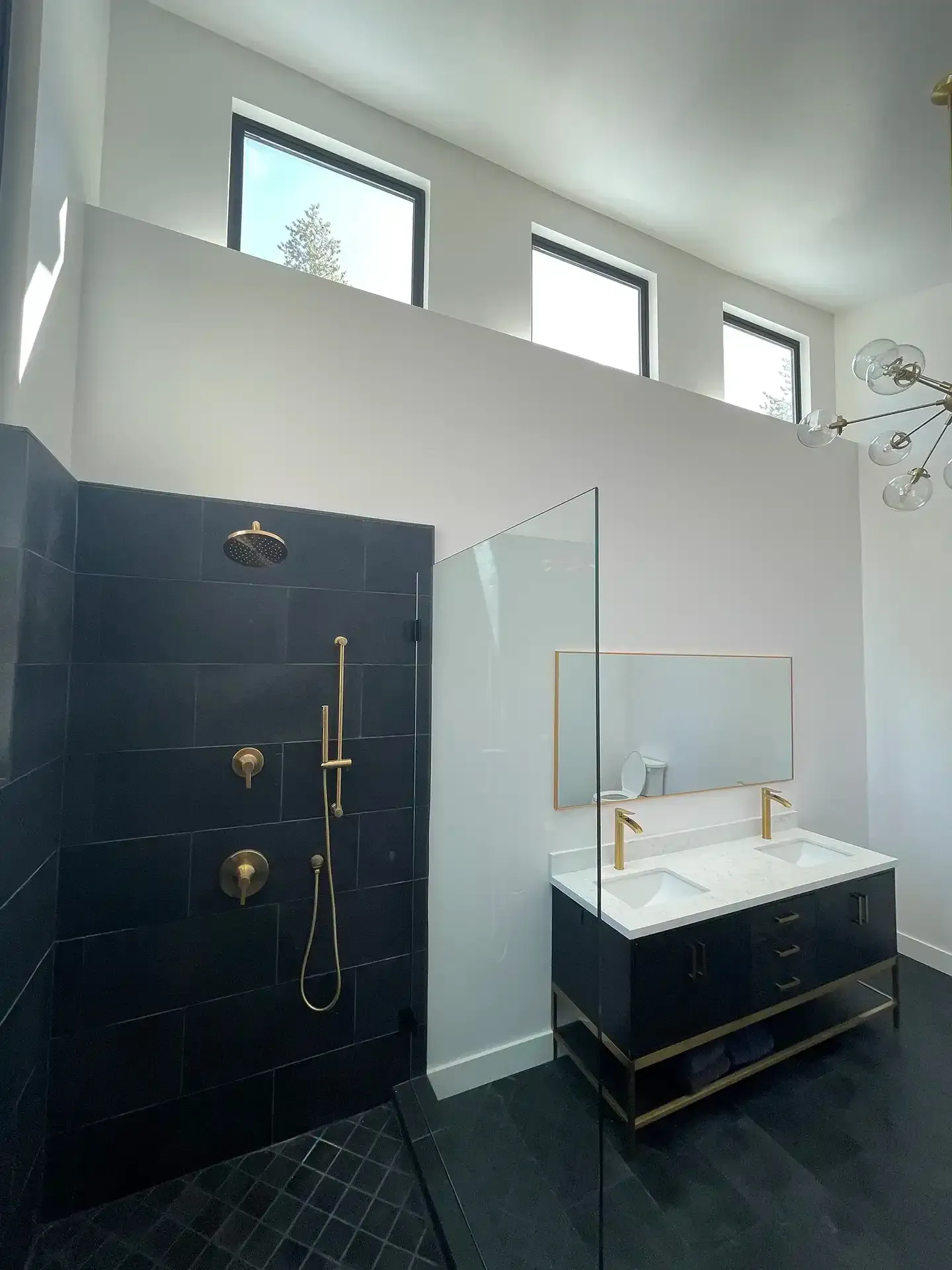 Modern bathroom with a dark tiled shower, a double vanity with gold fixtures, and three transom windows high on the wall.