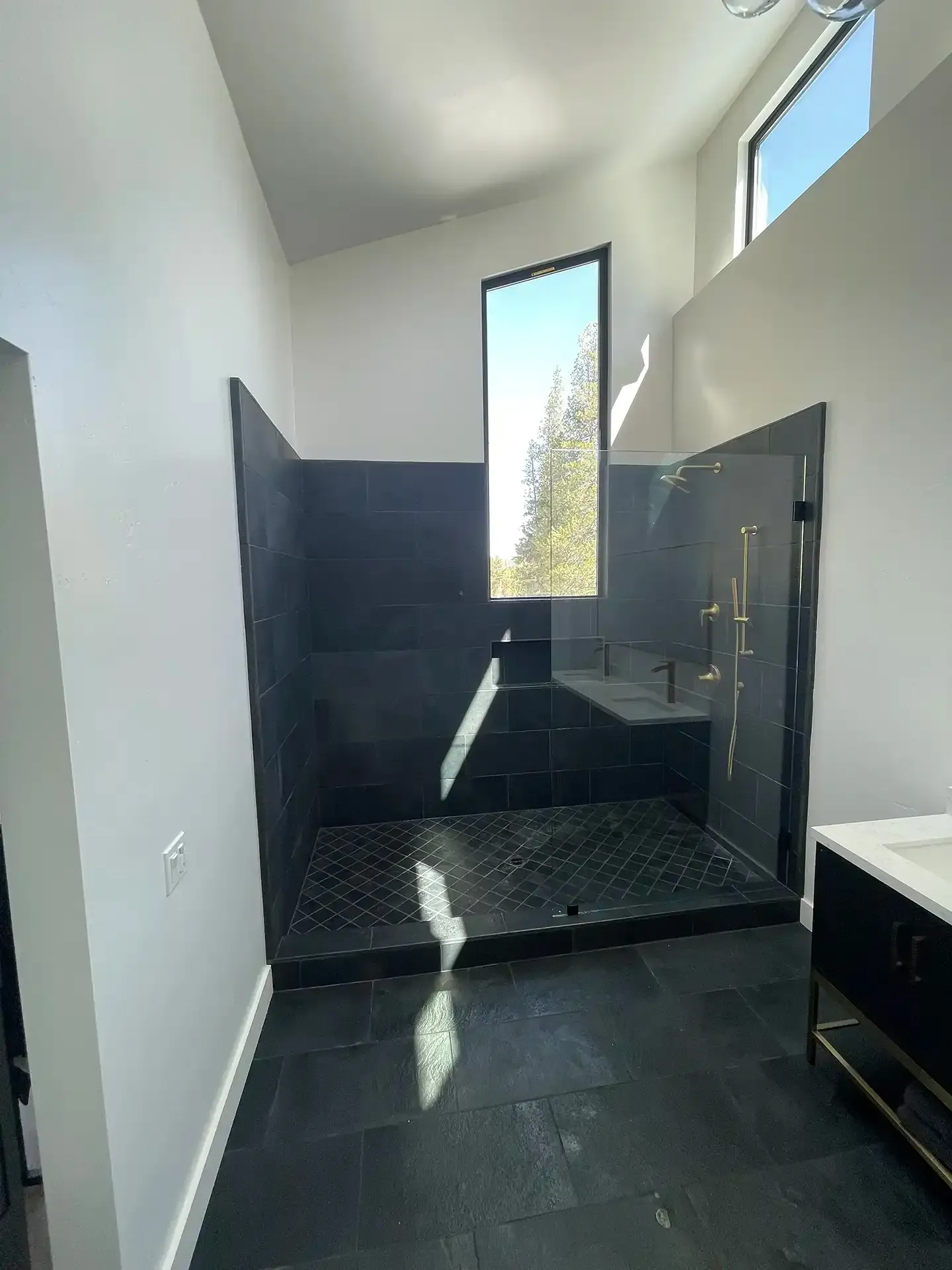 A modern bathroom with a black tile walk-in shower, glass door, and a tall, narrow window against white walls.