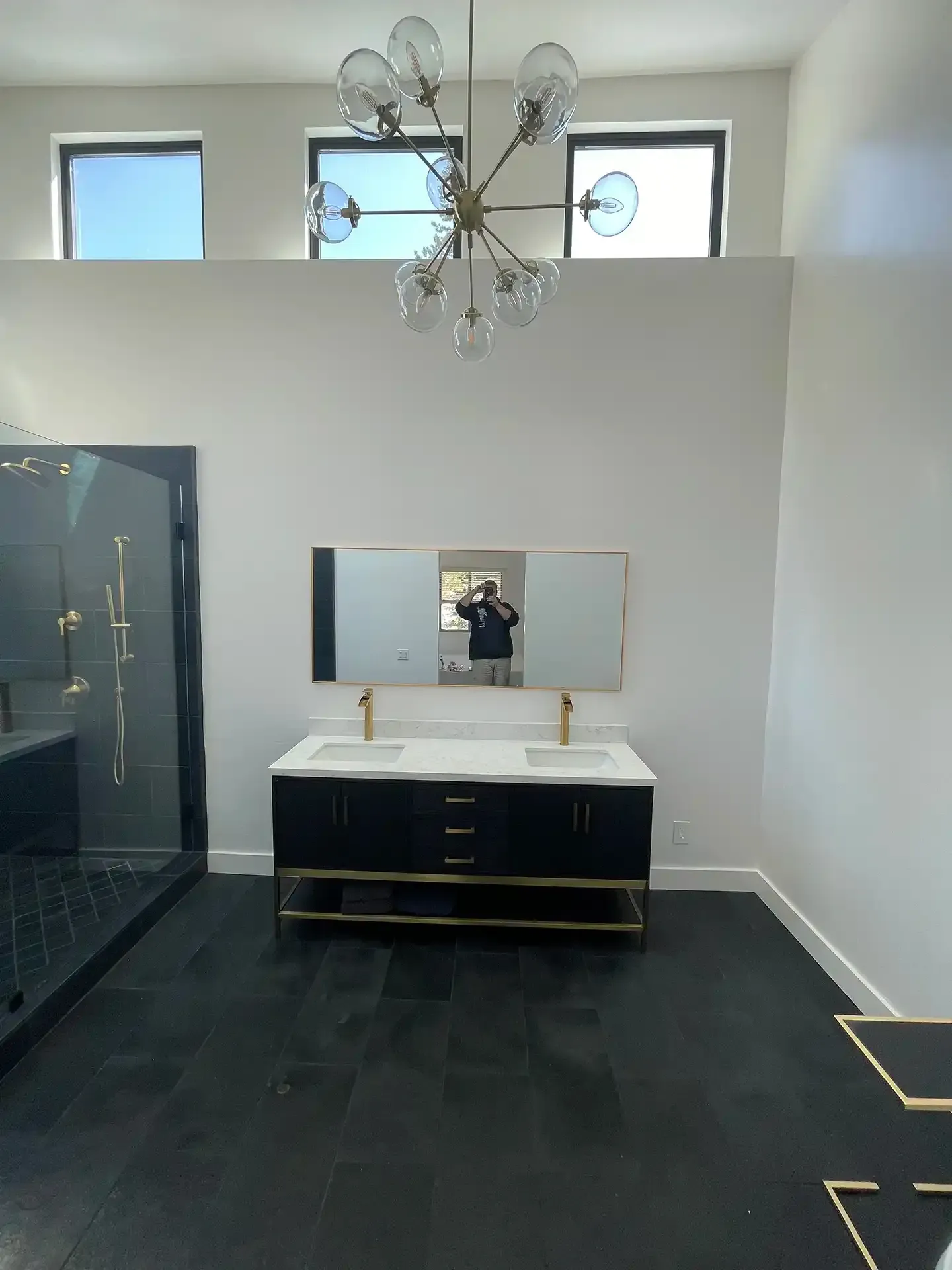 A modern bathroom with a black vanity, gold fixtures, a large mirror, dark tile floors, and a globe chandelier.