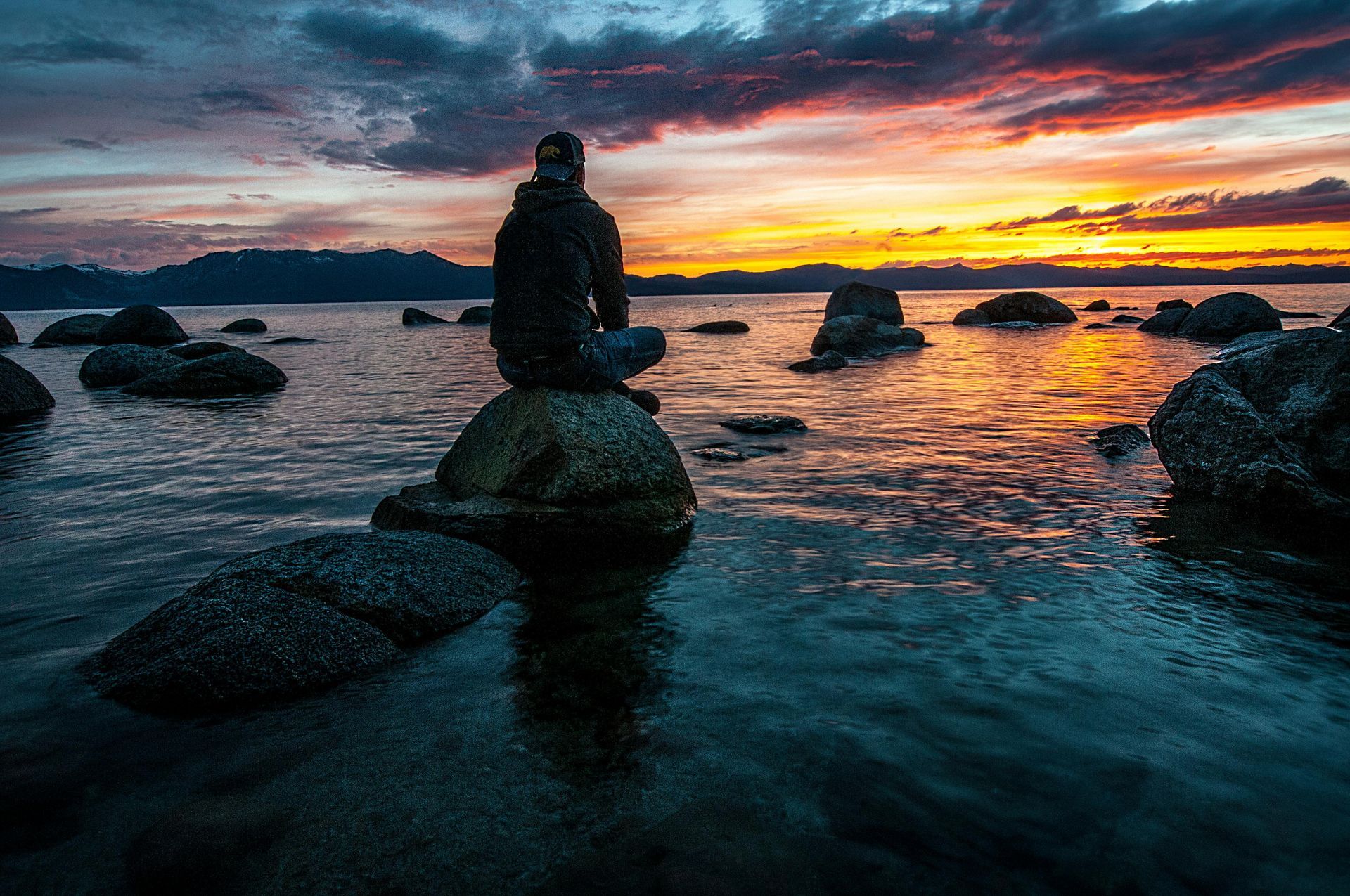 A person sits on a rock in a calm sea during a vibrant orange and purple sunset.