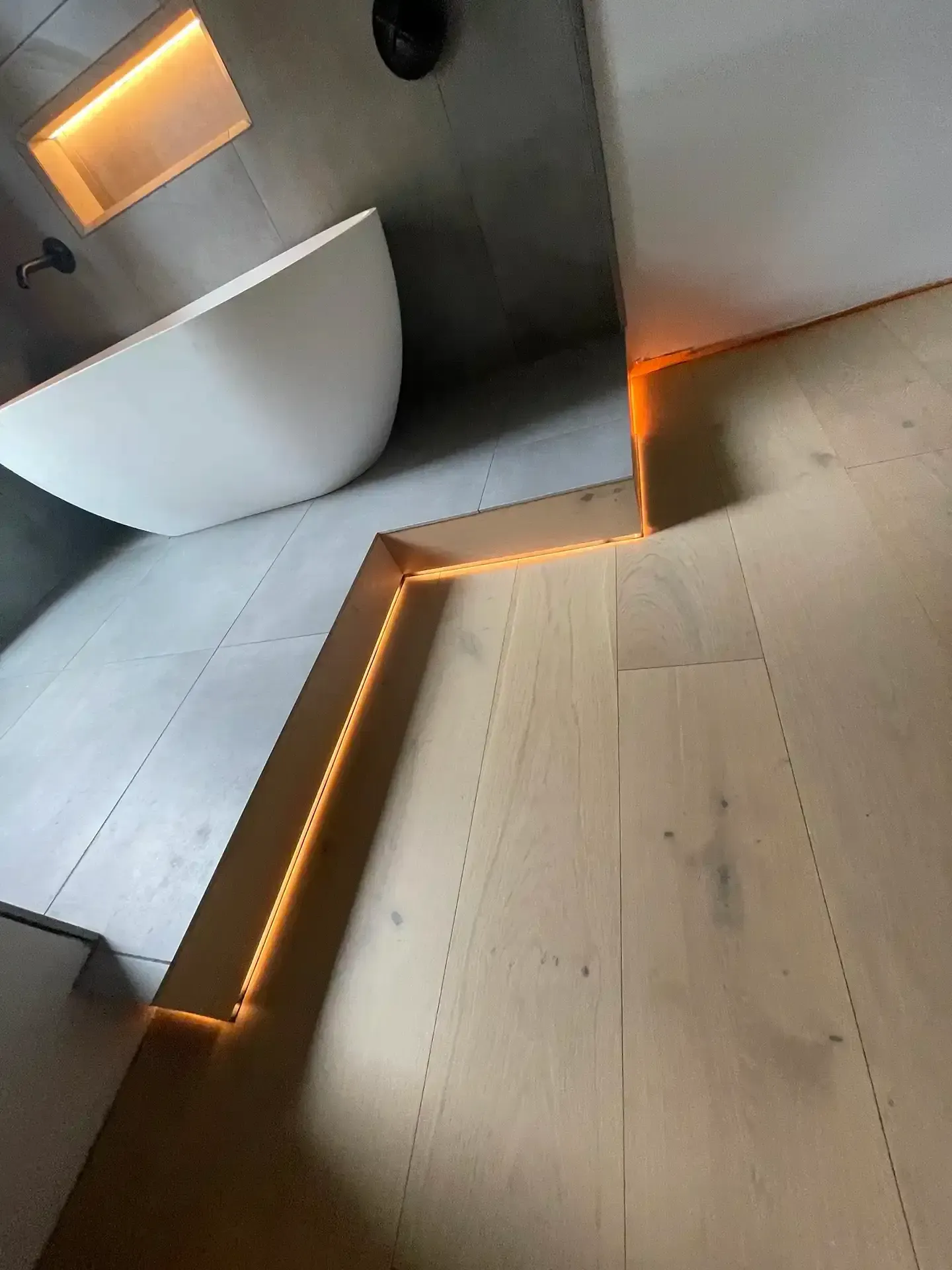 A modern bathroom featuring a white soaking tub on a raised, grey-tiled platform separated by integrated ambient lighting.