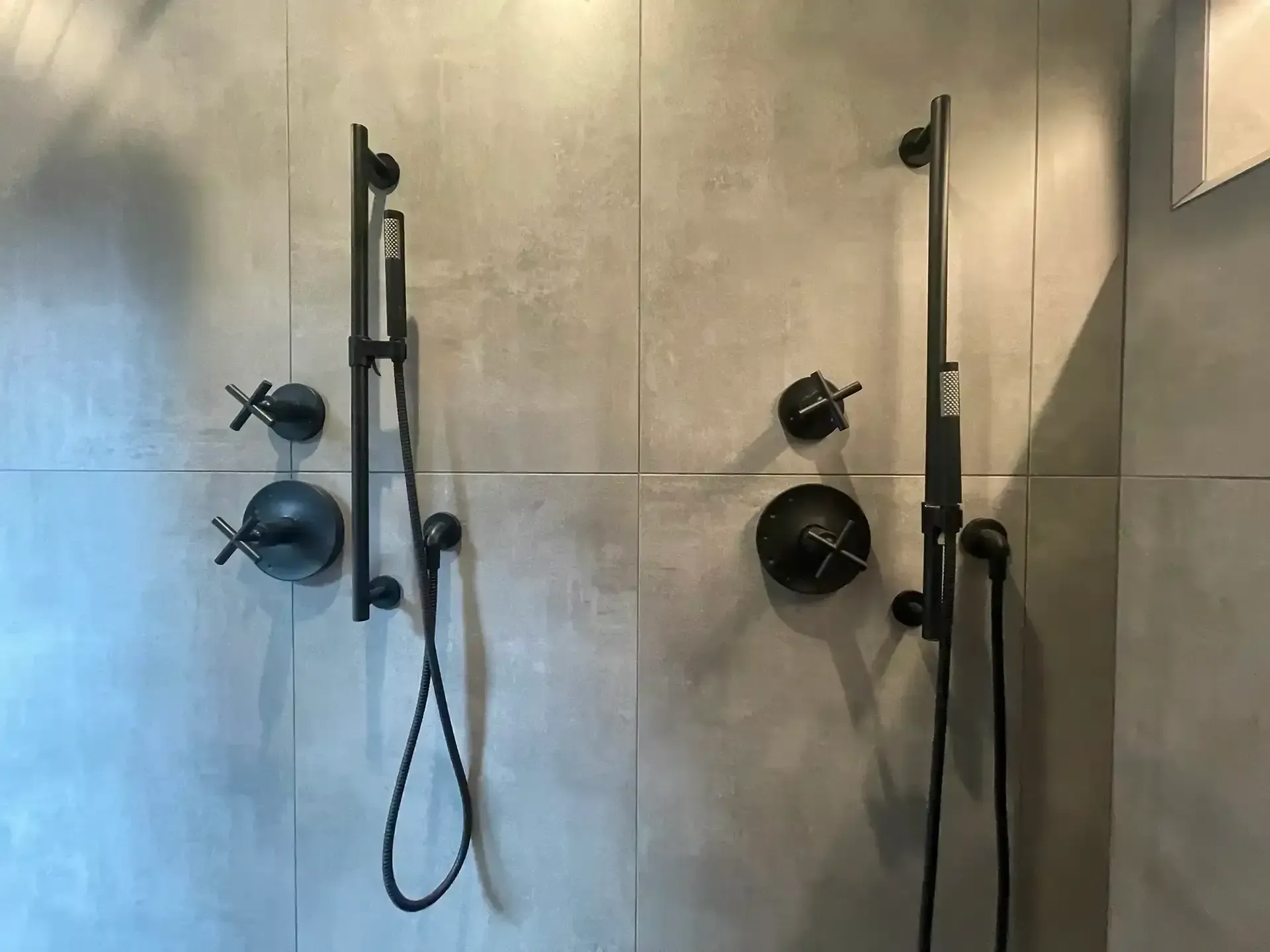 A shower wall with two sets of matte black hand showers, slide bars, and control knobs against large gray tile.