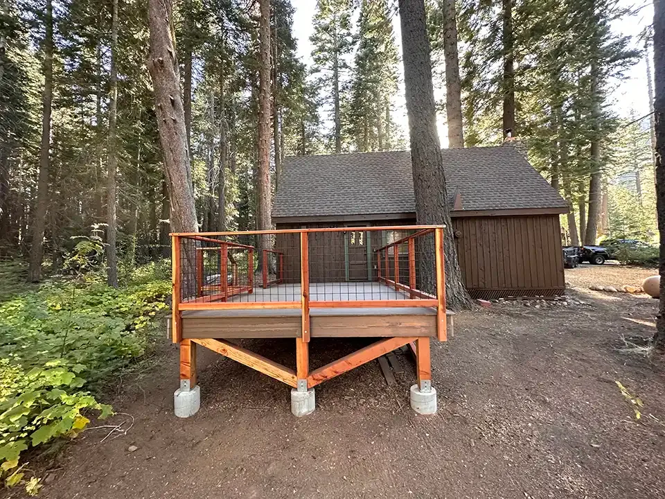 A wooden deck with a metal railing stands in a forest clearing in front of a small cabin nestled among pine trees.