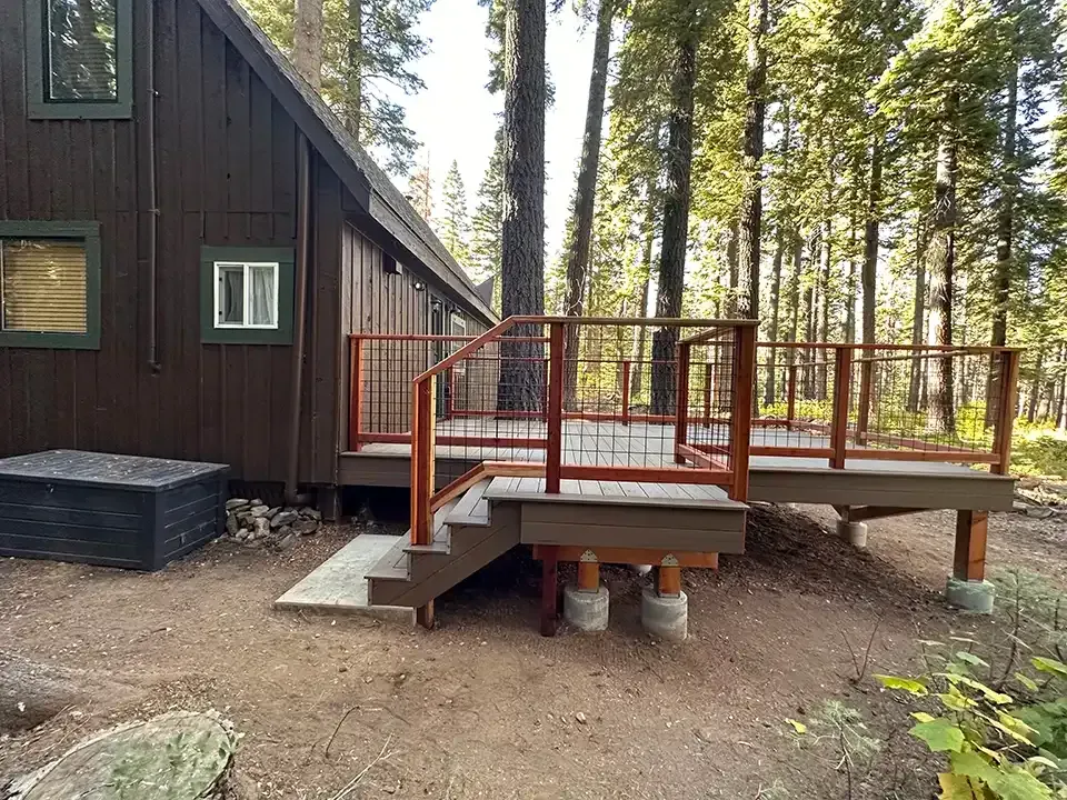 A dark brown wooden cabin with a modern elevated deck featuring cable railings and stairs, set among pine trees.