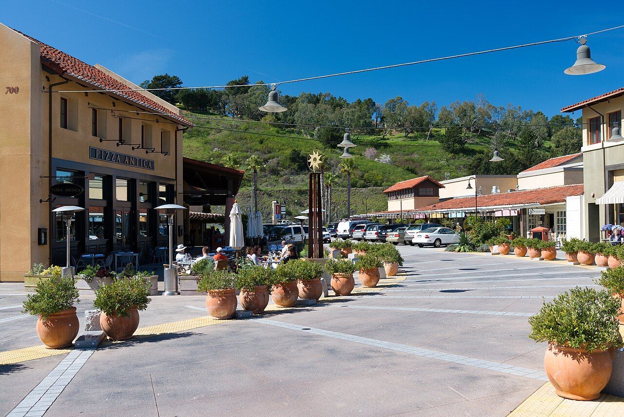 An outdoor plaza with terracotta-colored buildings, patio seating, and potted shrubs lining a paved walkway.