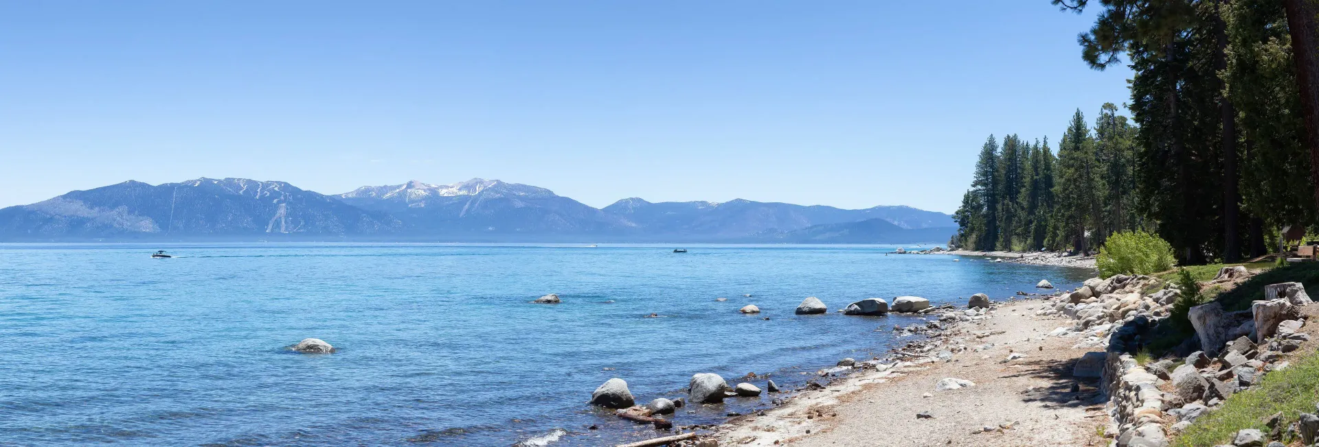 A clear blue lake with a rocky shoreline, pine trees on the right, and distant snow-capped mountains under a clear sky.