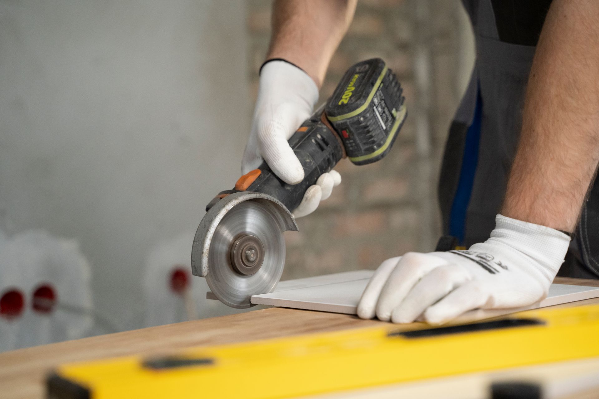 A person wearing white gloves uses a handheld angle grinder to cut a ceramic tile on a wooden workbench.