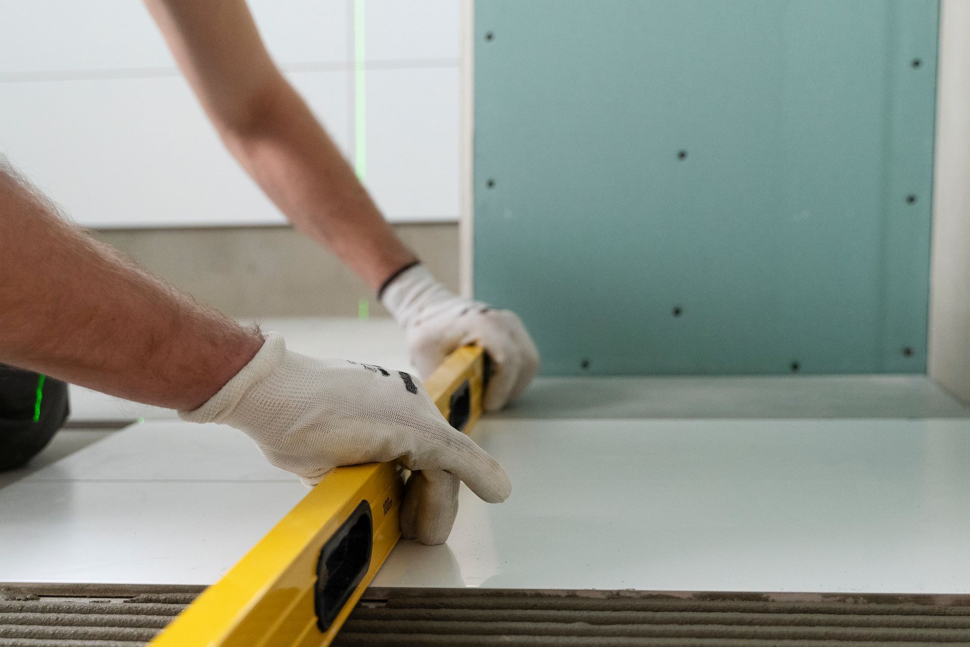 A person in white gloves uses a yellow spirit level to check the flatness of white floor tiles laid against a drywall wall.