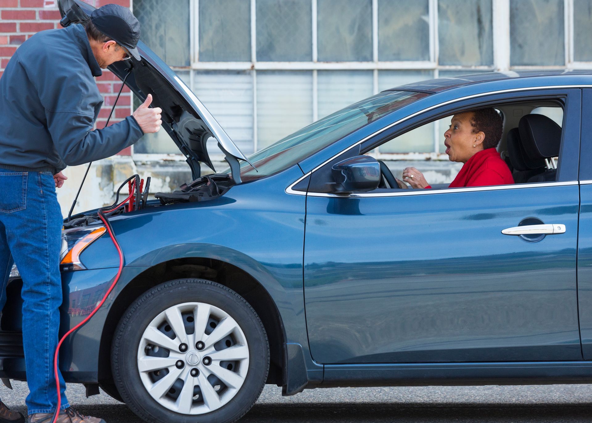 A towing technician providing a battery jump start for a woman’s car during roadside assistance.
