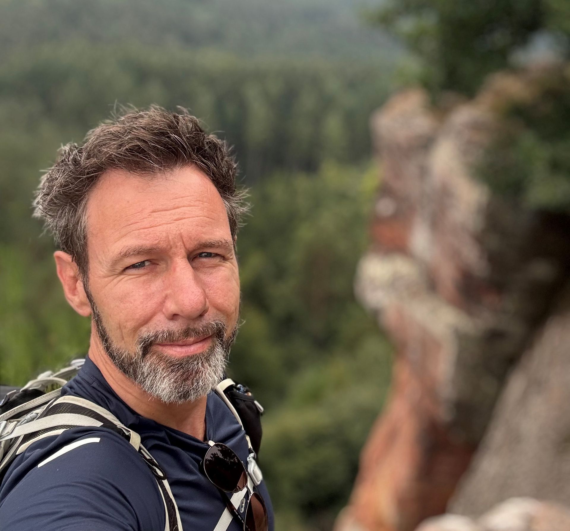 Een glimlachende man met een baard en een rugzak staat buiten, met op de achtergrond een wazig groen bos en rotsen.