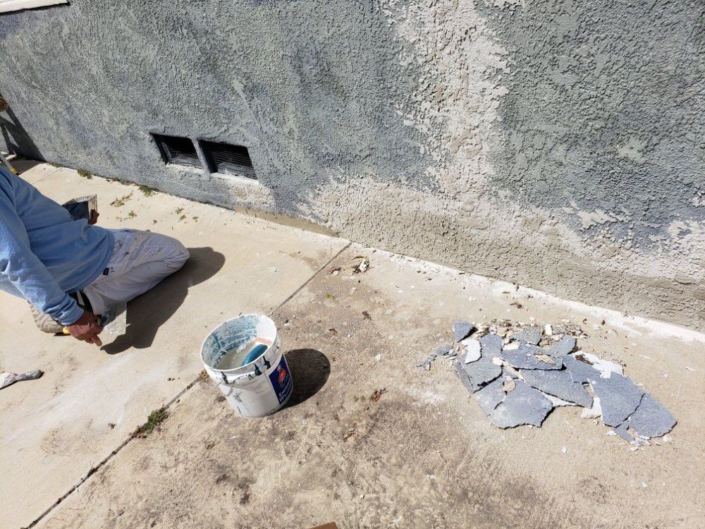 Person kneeling, repairing stucco on a building's exterior. Blue paint bucket, debris, and concrete ground visible.