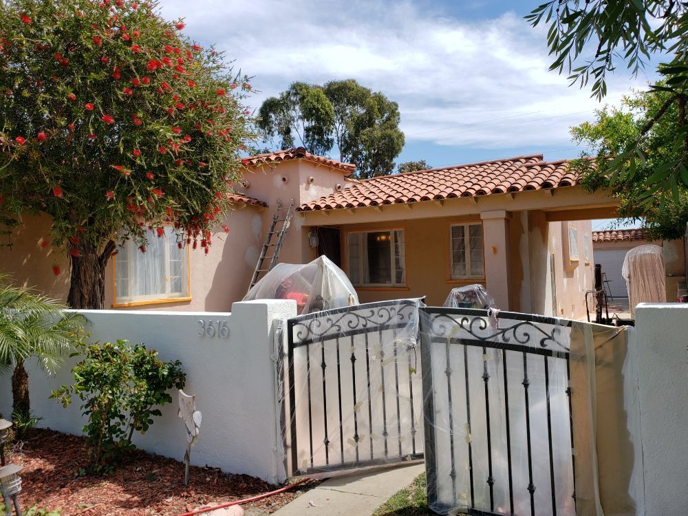 House being painted, behind a white wall with a black gate. Red roof and trees, bright sky.