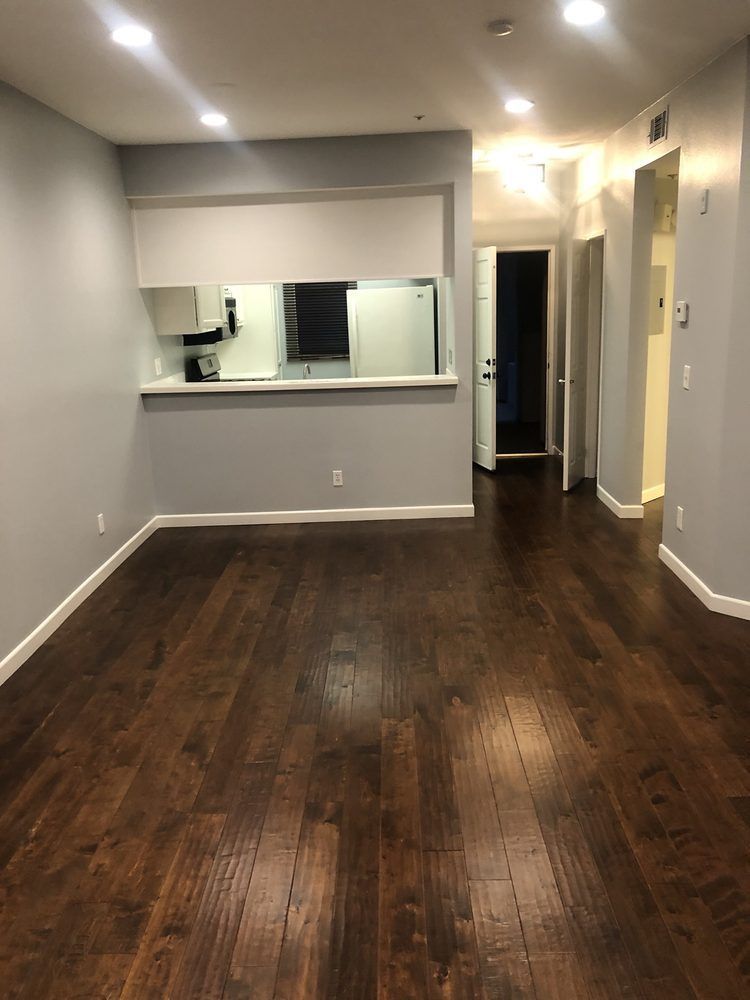 Empty, modern living space with dark wood floors, light gray walls, and open kitchen.