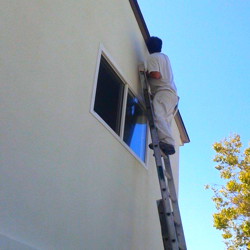 Person on a ladder painting the exterior of a two-story building. Clear blue sky.