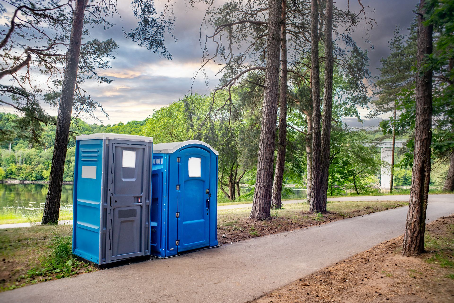 Two portable toilets are sitting next to each other on a sidewalk in the woods.