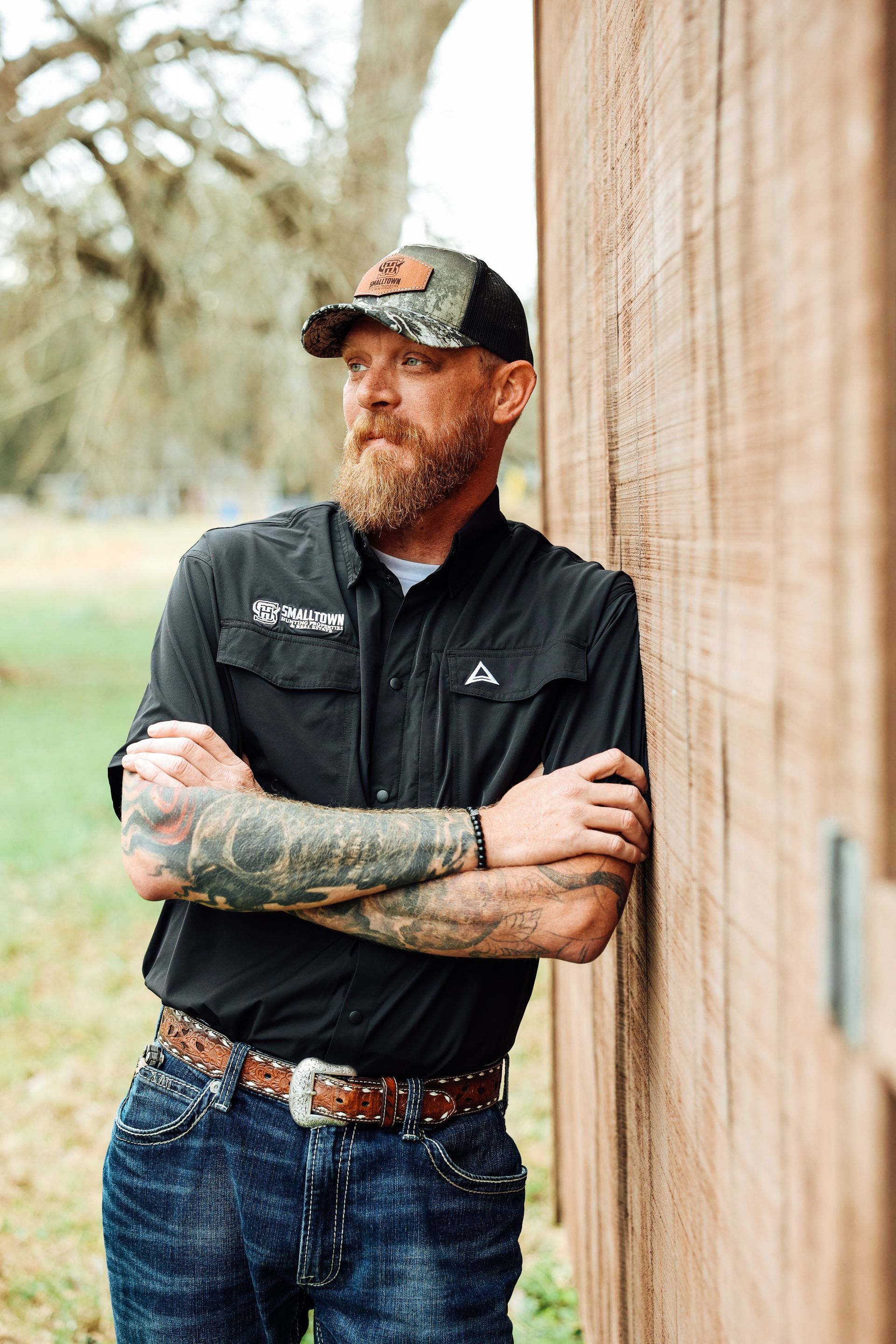 Man with beard and tattoos leans against a wooden wall, wearing a baseball cap and black shirt, arms crossed.