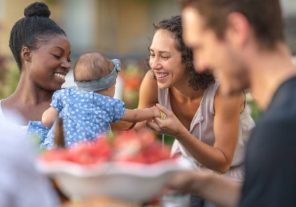 People smiling, reaching for baby in a blue dotted dress, outside near fruit.