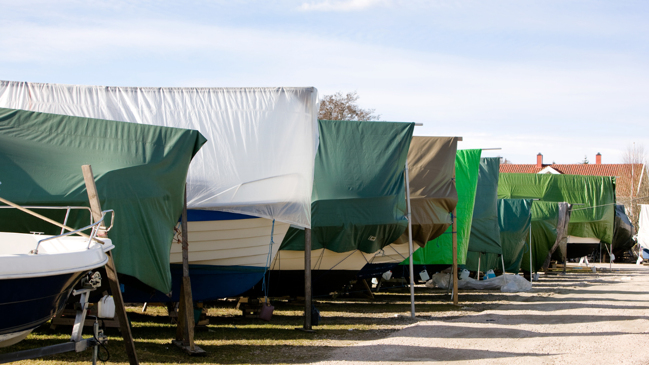 Boats covered in green and white tarps lined up in a row on a gravel lot under a blue sky.