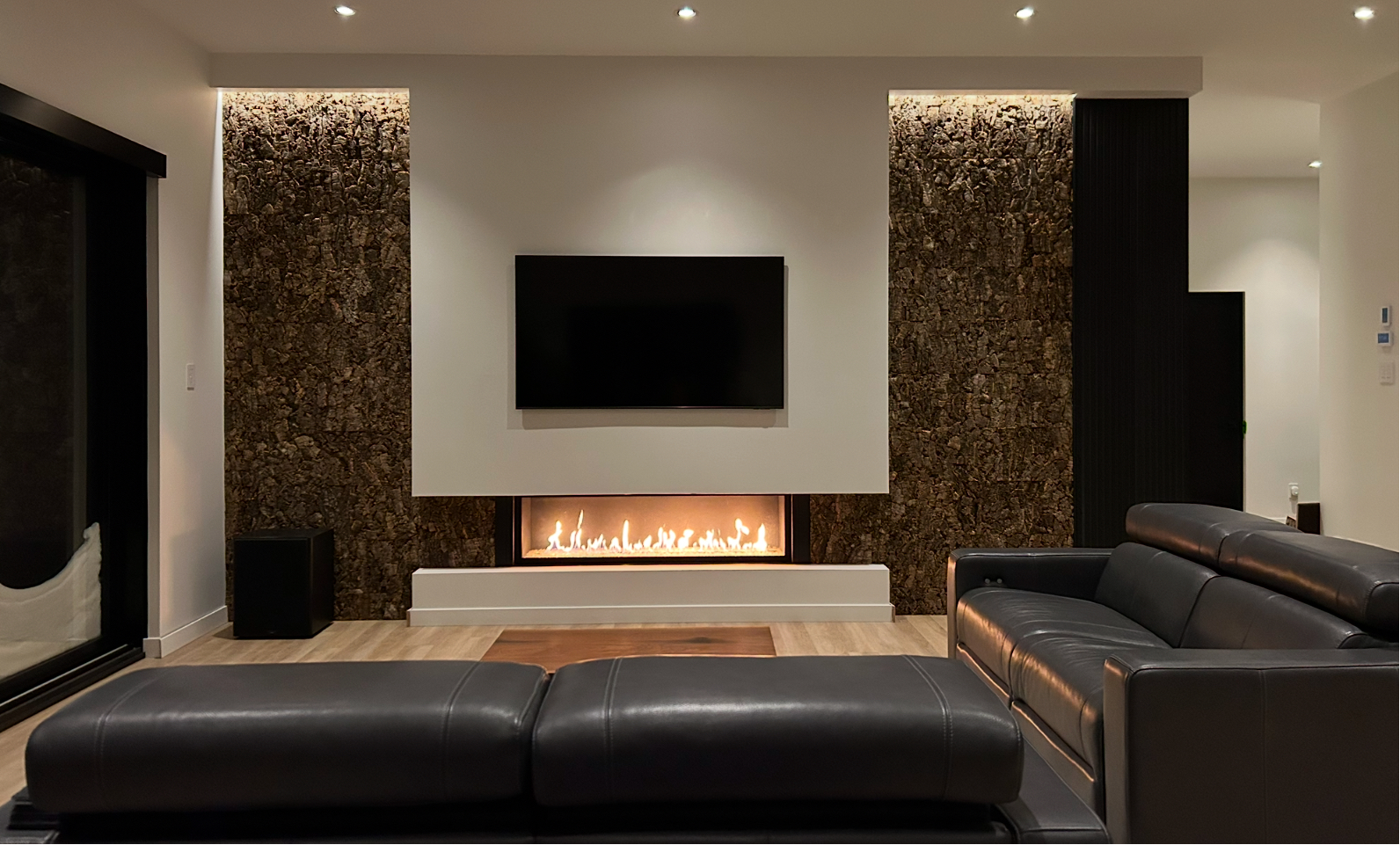 Salon avec cheminée, télévision et canapé en cuir noir. Le mur principal est orné de boiseries et d'éclairage encastré.