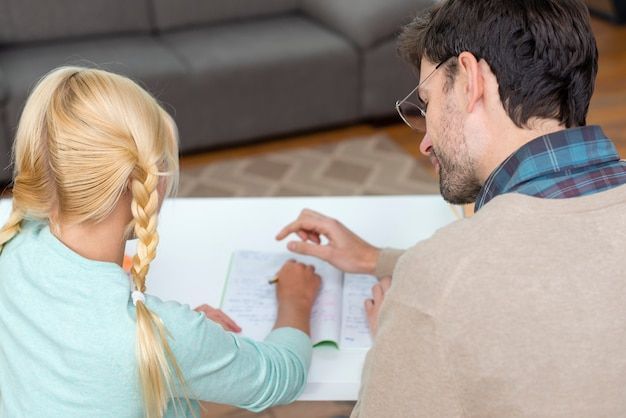 Blonde girl with pigtails writing at table, supervised by man with glasses, indoors.