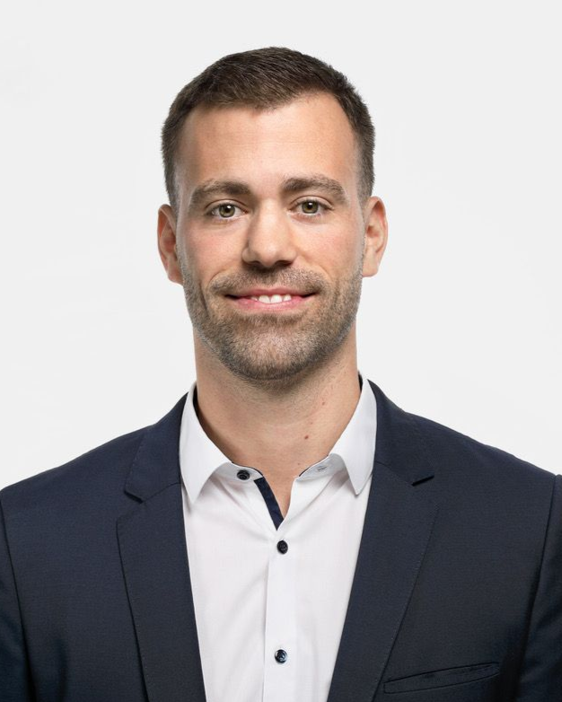 Ein Mann in einem dunkelblauen Blazer und einem weißen Hemd lächelt und schaut in die Kamera.