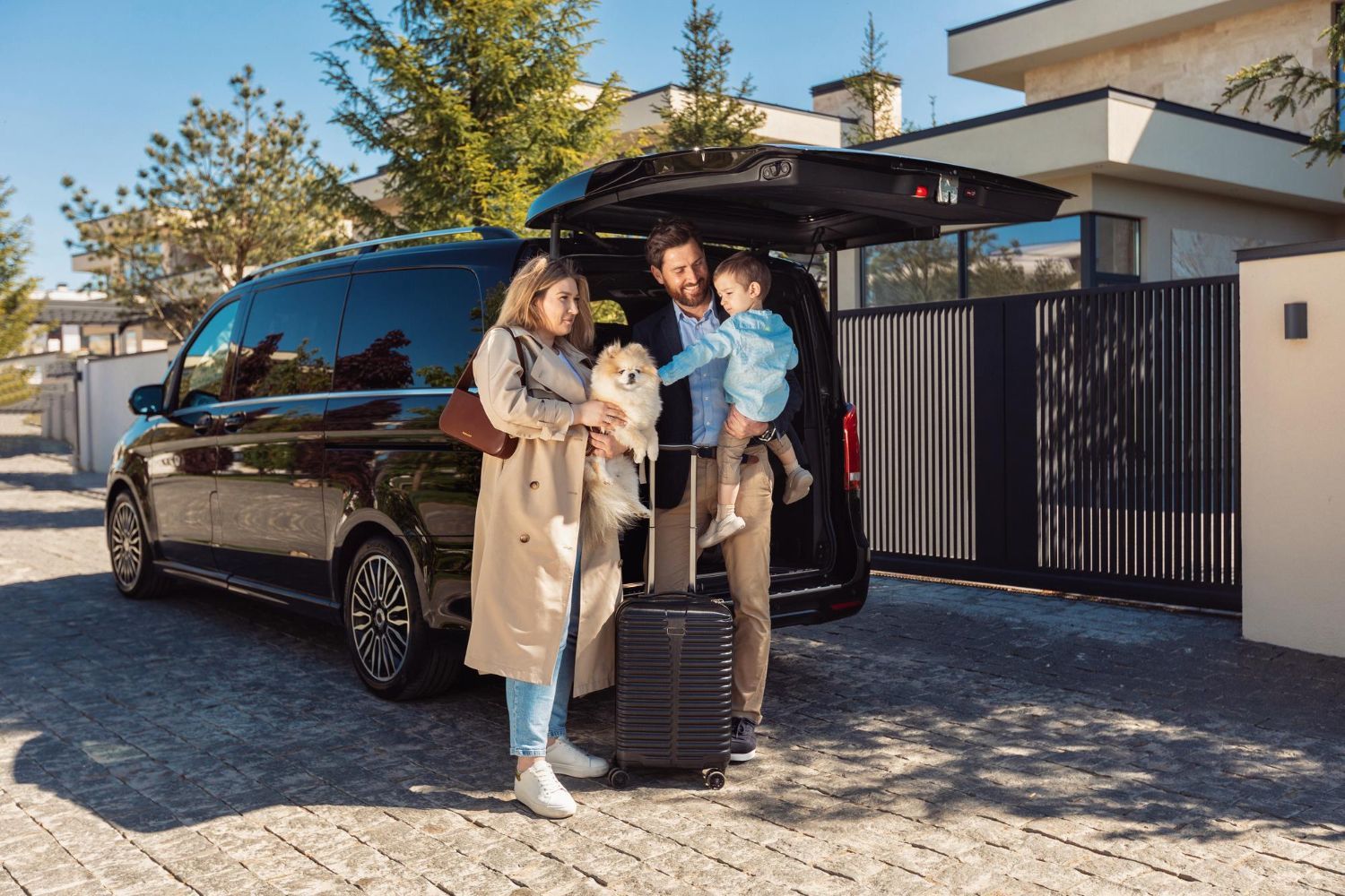 Family with luggage and small dog loading a black van outside a house.