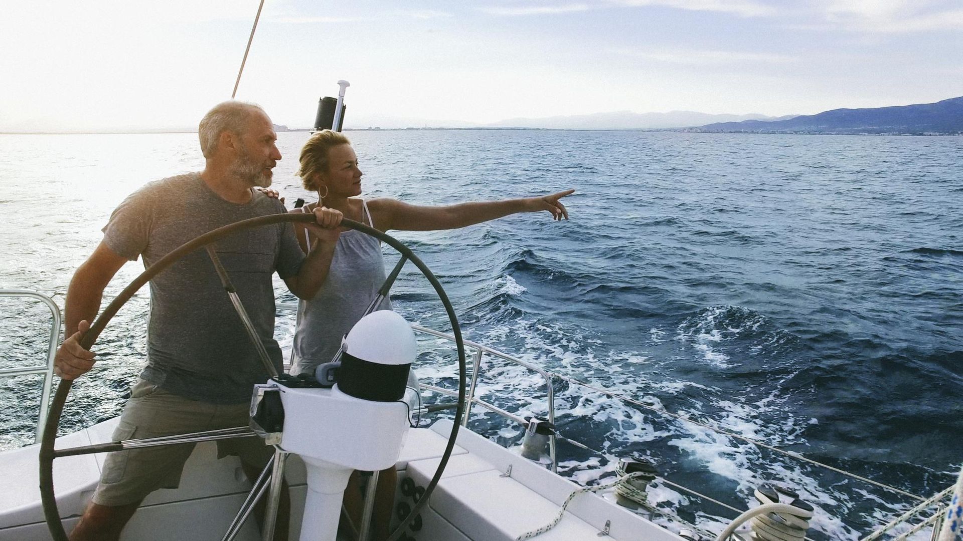 Man at the helm, woman points at horizon, both on a sailboat at sea.