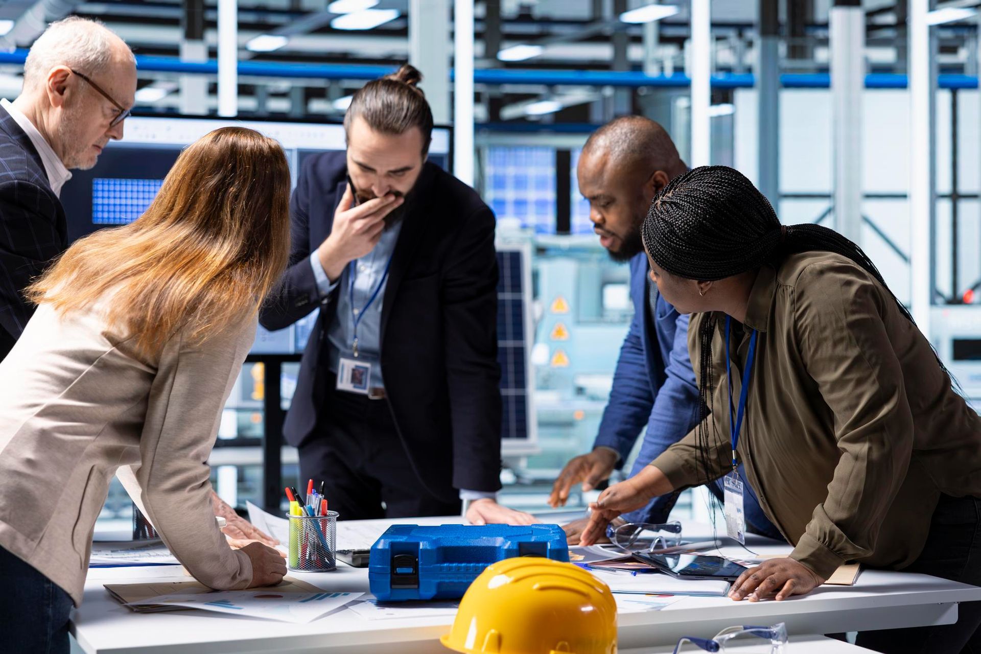 Team of people collaborating around a table with blueprints, discussing a project in a bright, industrial setting.